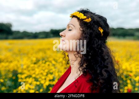 Vue latérale de la femme tendance dans la sundress rouge et avec couronne de fleur debout avec les yeux fermés sur le champ de floraison avec fleurs jaunes et rouges appréciant wa Banque D'Images