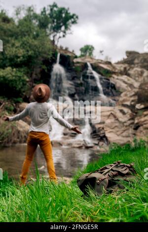 Vue arrière anonyme homme Backpacker dans le chapeau appréciant la vue de cascade en streaming depuis le roc dans la nature verdoyante Banque D'Images