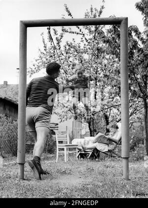 Père pousse l'enfant à bascule pendant que la mère lit en arrière-plan, 50s [traduction automatique] Banque D'Images