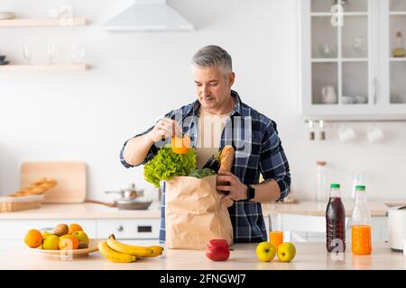 Concept de livraison de nourriture. Joyeux homme senior déballant le sac en papier avec l'épicerie commandée sur Internet Banque D'Images