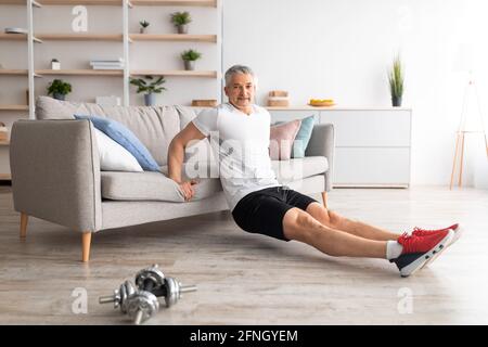 Entraînement à domicile pour rester en forme. Heureux homme mûr se penchant sur le canapé et faisant des exercices, en s'exerçant dans l'intérieur de salon Banque D'Images