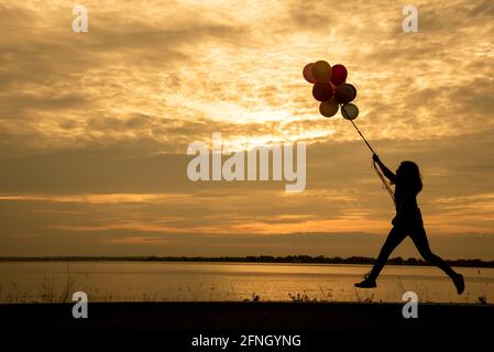 Silhouette femme tenant des ballons d'air debout et de voir le lac au coucher du soleil Banque D'Images