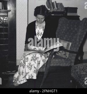 Années 1950, historique, une dame portant une longue robe à motif fleuri assise dans une chaise en bois dans une salle de lecture d'un magazine, Angleterre, Royaume-Uni. Banque D'Images