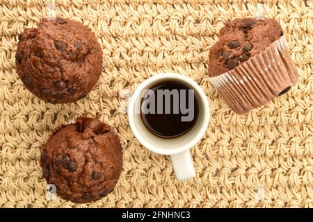 Trois muffins au chocolat et une tasse blanche avec café, sur un tapis de paille, vue de dessus. Banque D'Images