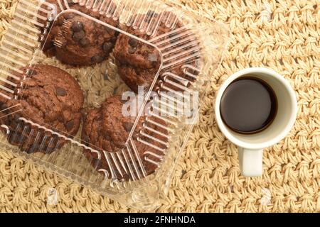 Plusieurs muffins au chocolat dans un récipient en plastique avec une tasse de café, gros plan, sur un tapis de paille, vue de dessus. Banque D'Images