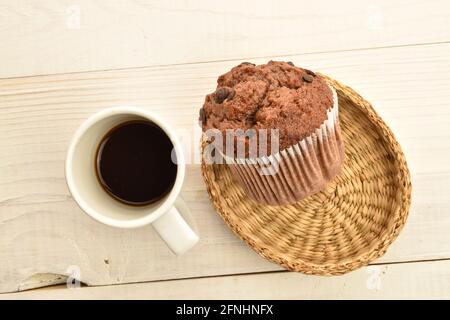 Un muffin au chocolat sur une assiette de paille avec une tasse de café, gros plan, sur une table en bois blanc, vue du dessus. Banque D'Images