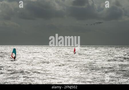 Image stupéfiante de deux planches à voile prenant le soleil, l'une avec une voile bleue et l'autre avec rouge, sur une mer argentée avec un troupeau d'oiseaux contre un ciel sombre Banque D'Images