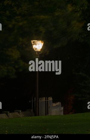 Lampe de rue unique de style ancien éclairée la nuit sur fond noir et éclairant un mur en pierre et une clôture en fer avec un premier plan gazonné Banque D'Images