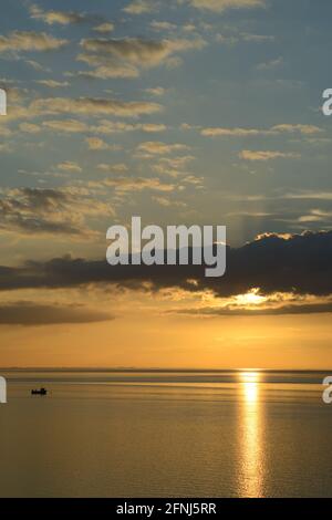 Image couleur du soleil coulant derrière le nuage et l'éclairage dans un éclat doré à la fois la mer et le ciel avec un bateau de pêche éloigné en silhouette Banque D'Images