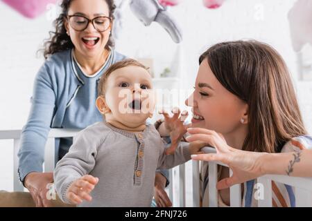 une mère souriante qui fait des gestes près d'un enfant émerveillé dans un lit de bébé et un ami afro-américain enthousiaste Banque D'Images