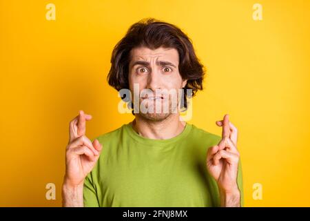 Photo de la lèvre de morsure de jeune homme inquiet montrant les doigts croisés porter un t-shirt vert isolé de couleur jaune arrière-plan Banque D'Images