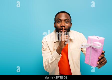 un jeune homme afro-américain tient une boîte cadeau tout en montrant la huche signe isolé sur bleu Banque D'Images