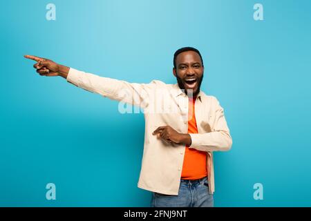 rire d'un homme afro-américain regardant la caméra et pointant du côté avec le doigt sur fond bleu Banque D'Images