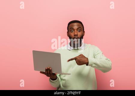 surpris afro-américain homme pointant vers l'ordinateur portable sur fond rose Banque D'Images