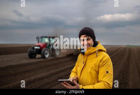 Portrait d'un beau fermier avec une tablette debout devant le tracteur herme dans le champ en arrière-plan Banque D'Images