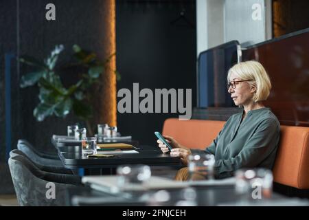 Femme mûre pensive sérieuse avec cheveux blonds assis à la table dans un café à la mode et en vérifiant les notes téléphoniques Banque D'Images