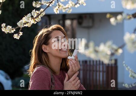 Jolie jeune femme utilisant la pompe d'asthme devant l'arbre en fleur au printemps. Attaque contre les allergies saisonnières Banque D'Images