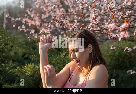 Jolie jeune femme se grattant le bras à côté de l'arbre en fleurs dans le parc au printemps. Rougeur et démangeaisons de la peau comme réaction allergique Banque D'Images