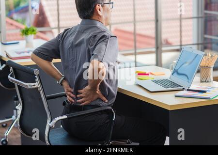 Un homme d'affaires asiatique expérimenté travaillant avec un ordinateur portable a un problème avec les maux de dos. Le vieil homme ressent de la douleur après s'être assis longtemps à son bureau, Healthc Banque D'Images