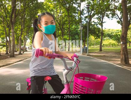 Petite fille asiatique portant un masque ou un masque facial pour prévenir le coronavirus, assise sur un vélo rose dans le parc de la Thaïlande. Activités de plein air Banque D'Images