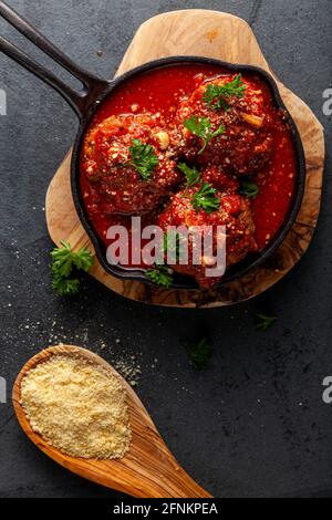 Vue rapprochée de trois grandes boulettes de viande traditionnelles italiennes dans une sauce tomate en mélange dans une casserole en fonte. La poêle repose sur des feuilles de bois et de persil Banque D'Images