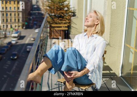 Le matin. Belle femme adulte rêveuse des années 60 dans une tenue décontractée se détendre sur le balcon à la maison, commencer la journée avec le café. Bonne femme âgée assise sur la terrasse et regardant la rue ensoleillée Banque D'Images