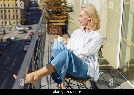 Le matin. Belle femme adulte rêveuse des années 60 dans une tenue décontractée se détendre sur le balcon à la maison, commencer la journée avec le café. Bonne femme âgée assise sur la terrasse et regardant la rue ensoleillée Banque D'Images
