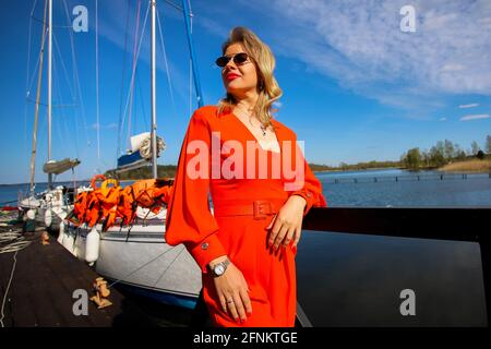 Belle femme dans le costume rouge vif sur le fond du yacht. La blonde en lunettes de soleil sur le fond du yacht et le ciel bleu. Banque D'Images