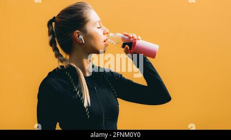 Jolie fille caucasienne sur fond jaune isolé avec casque sans fil en costume de sport faire de l'entraînement, courir et boire de l'eau Banque D'Images