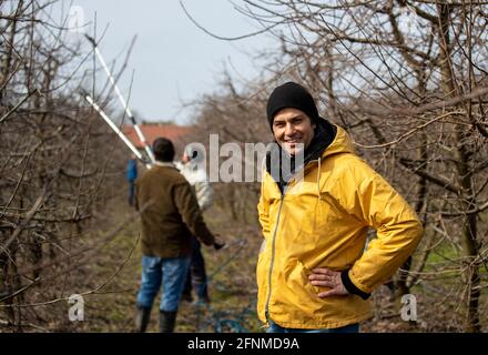 Portrait d'un beau fermier debout dans un verger en hiver pendant que les ouvriers élagage les arbres fruitiers en arrière-plan Banque D'Images