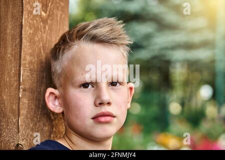 Joli garçon blond avec une coiffure courte s'appuie sur une clôture en bois poser pour caméra dans vert ensoleillé été ville parc extrême gros plan Banque D'Images