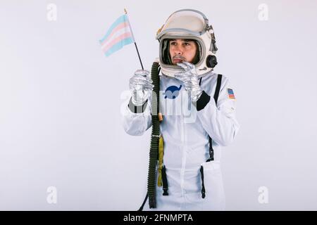 Cocosmonaute mâle transsexuel gay avec un geste sérieux en combinaison spatiale et casque, tenant le drapeau transgenre, sur fond blanc. Banque D'Images