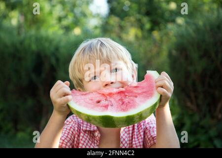 un petit garçon mignon avec l'appétit mange un grand morceau de pastèque. Délicieux fruits rouges préférés pour l'été. Banque D'Images