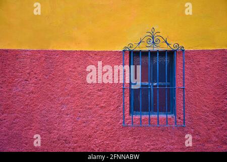 Façade de maison coloniale avec un mur texturé rose et ocre et une fenêtre avec des grilles en fer artisanales bleues à San Miguel de Allende, Mexique. Banque D'Images