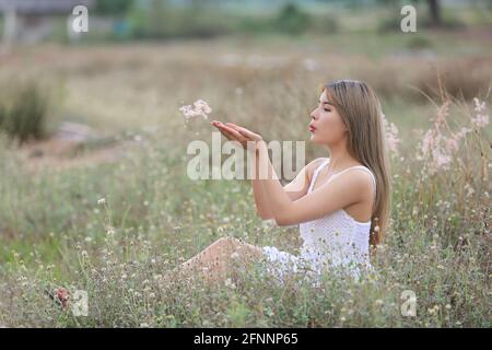 Une belle femme avec de longs cheveux dans une robe assise sur un pré et des fleurs dans la lumière du soir. Banque D'Images