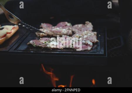 L'homme utilise une louche en acier pour tourner un col de porc chargé avec du basilic, du sel, du poivre et de l'huile d'un côté à l'autre. Barbecue sur un sapin extérieur féroce Banque D'Images