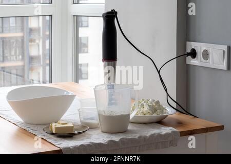 Un bol blanc, un mélangeur à immersion, branché dans la prise électrique, des récipients avec de la farine, du beurre, du sucre, du fromage cottage se tiennent sur une table près de la fenêtre. T Banque D'Images