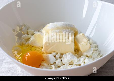 Fromage cottage, beurre, œuf et sucre dans un bol blanc - ingrédients préparés pour le mélange. Le processus de fabrication de petits pains ou de croissants caillé Banque D'Images