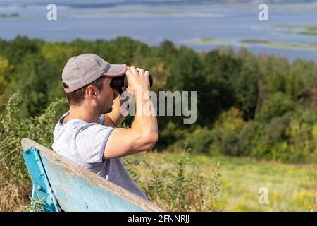 Un homme regarde à travers des jumelles une vue panoramique sur une rivière large tout en étant assis sur un vieux banc en bois sur une colline. Vue latérale Banque D'Images