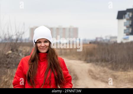 Souriant belle jeune femme avec de longs cheveux dans un blanc bonnet tricoté et veste rouge avec capuche la rue par une journée nuageux contre un arrière flou Banque D'Images