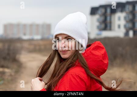 Jeune femme souriante avec de longs cheveux en bonnet blanc tricoté et une veste rouge avec capuche et vue sur l'épaule la rue le jour nuageux contre flou Banque D'Images