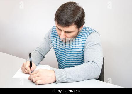 Beau homme attentionné dans un chandail rayé se trouve à la table et écrit avec un stylo sur papier Banque D'Images