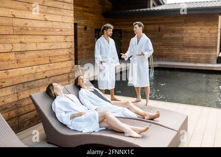 Deux amies sont allongé dans des peignoirs sur des chaises longues à l'extérieur après des soins au spa et au sauna, tandis que deux hommes se tiennent côte à côte et parlent. Bien-être et loisirs ensemble. Banque D'Images