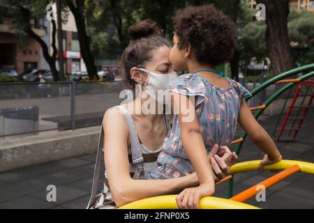 Jolie fille embrassant sa mère avec un masque facial à l'aire de jeux extérieur Banque D'Images