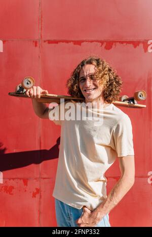 portrait vertical d'un jeune homme portant un skateboard sur cet épaule. Il a des cheveux longs bouclés rouges et des robes décontractées. Arrière-plan rouge avec shado profond Banque D'Images