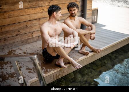 Des amis de sexe masculin assis sur la terrasse près du lac dans le complexe thermal et parlant. Détendez-vous et amusez-vous dans le centre de bien-être. Banque D'Images