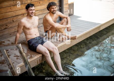 Des amis de sexe masculin assis sur la terrasse près du lac dans le complexe thermal et parlant. Détendez-vous et amusez-vous dans le centre de bien-être. Banque D'Images