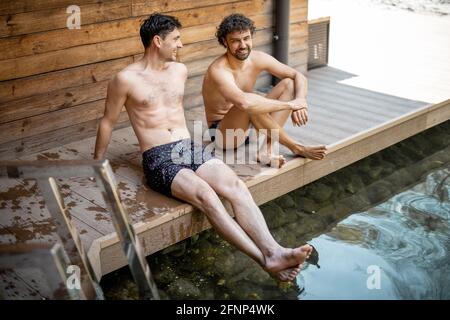 Des amis de sexe masculin assis sur la terrasse près du lac dans le complexe thermal et parlant. Détendez-vous et amusez-vous dans le centre de bien-être. Banque D'Images