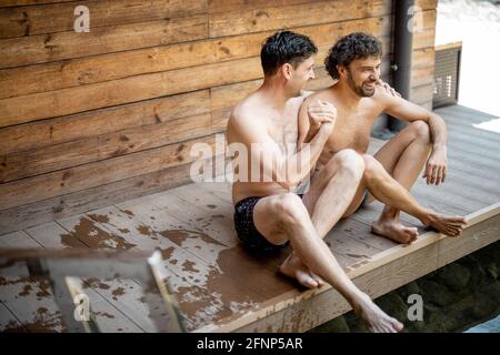 Des amis masculins assis sur la terrasse près du lac dans le complexe de spa tout en parlant et en tenant les mains. Détendez-vous et amusez-vous dans le centre de bien-être. Banque D'Images