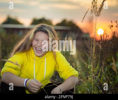 Une jeune fille avec de longs cheveux sur un pré avec de l'herbe haute dans les rayons du soleil couchant. Cheveux blonds. Veste jaune. Banque D'Images